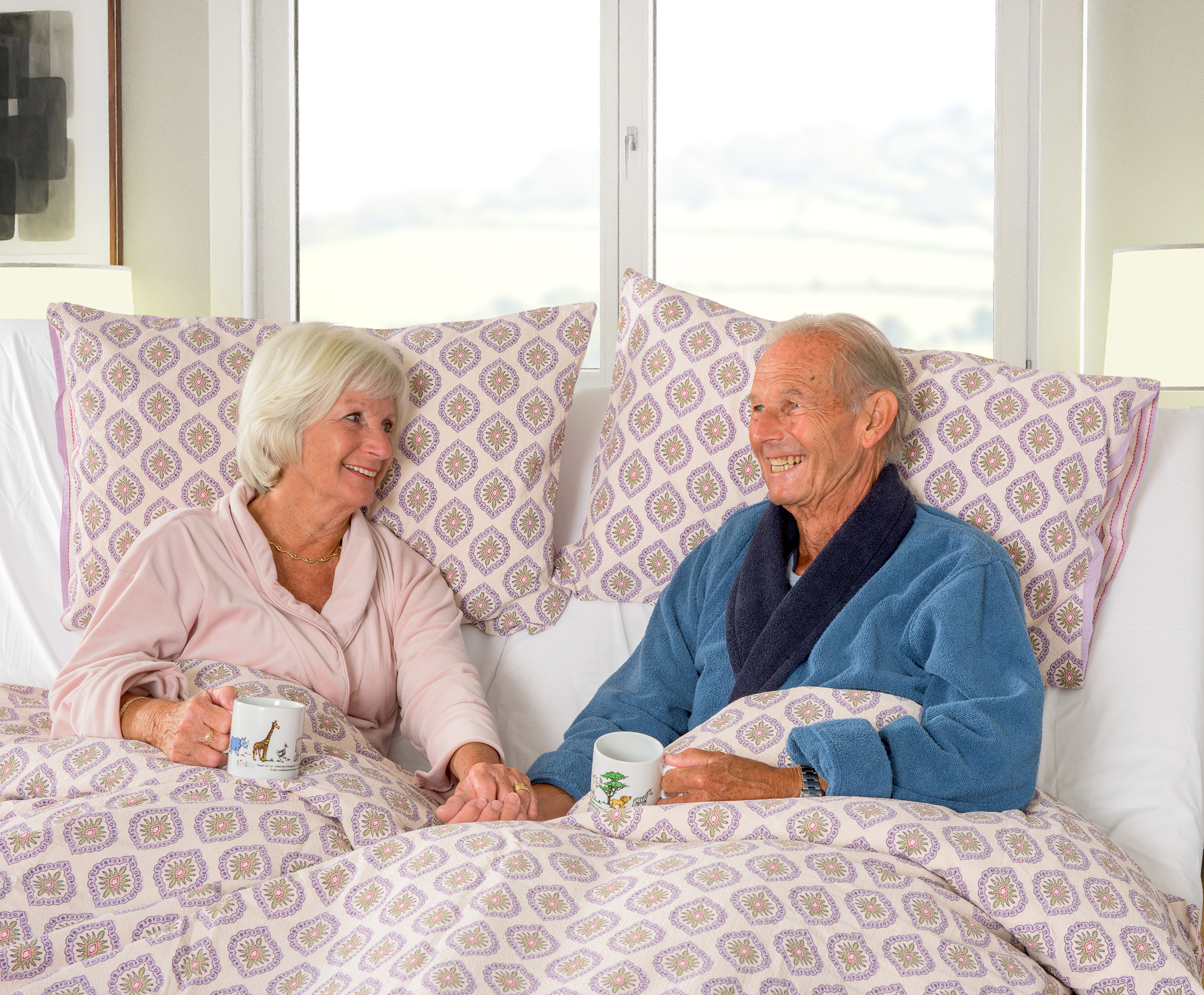 Couple of elderly people laying in bed with cups, holding hands and talking